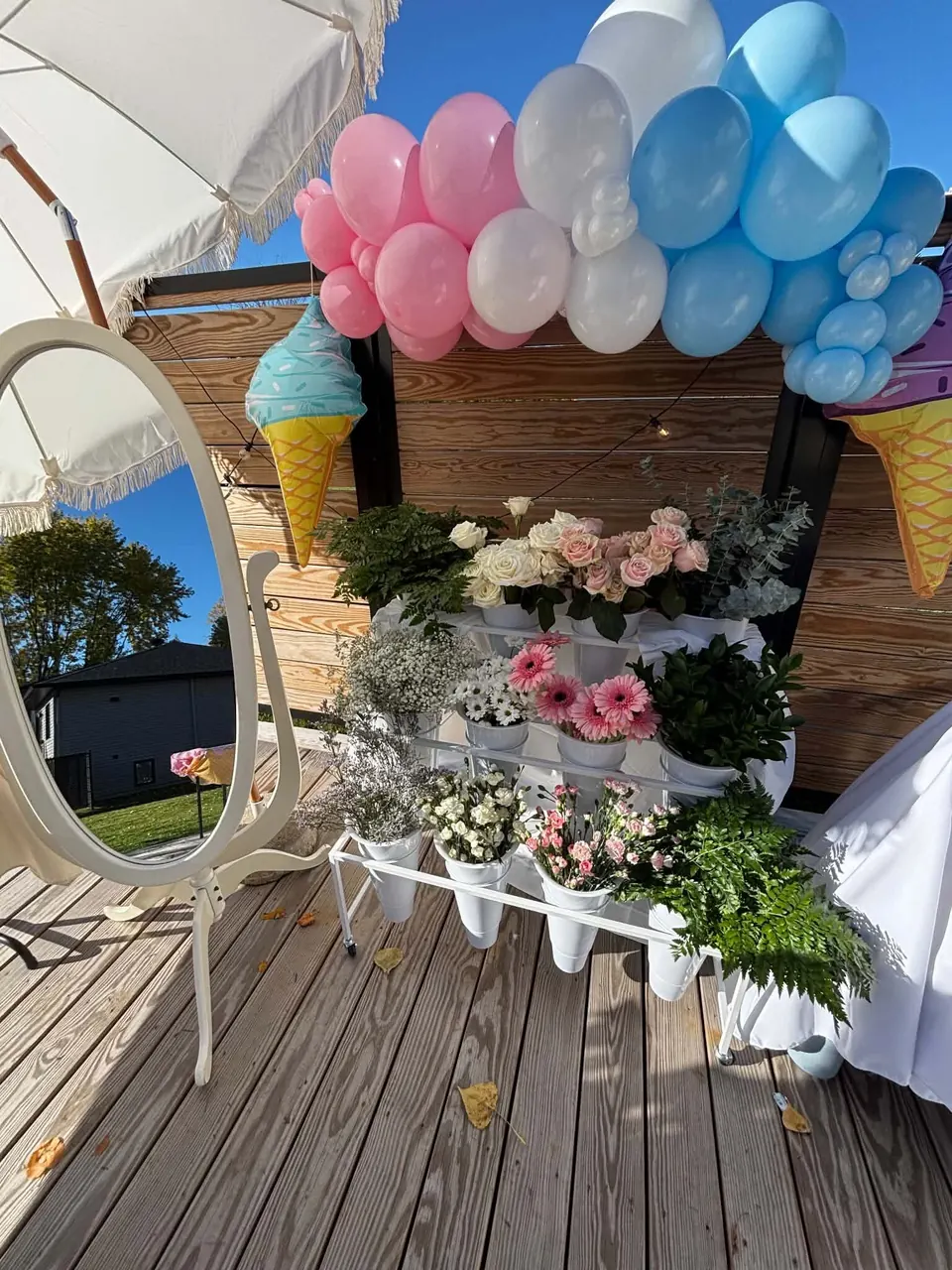 Bar à fleurs blanc sous un parasol à franges, miroir ovale et arche de ballons rose, blanc et bleu sur une terrasse en bois