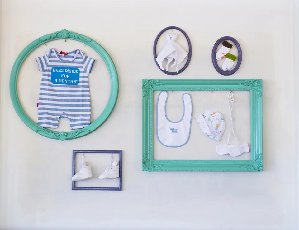 Cadres vides peints en turquoise et bleu marine accrochés au mur, avec une grenouillère, un bavoir et des chaussons à l’intérieur