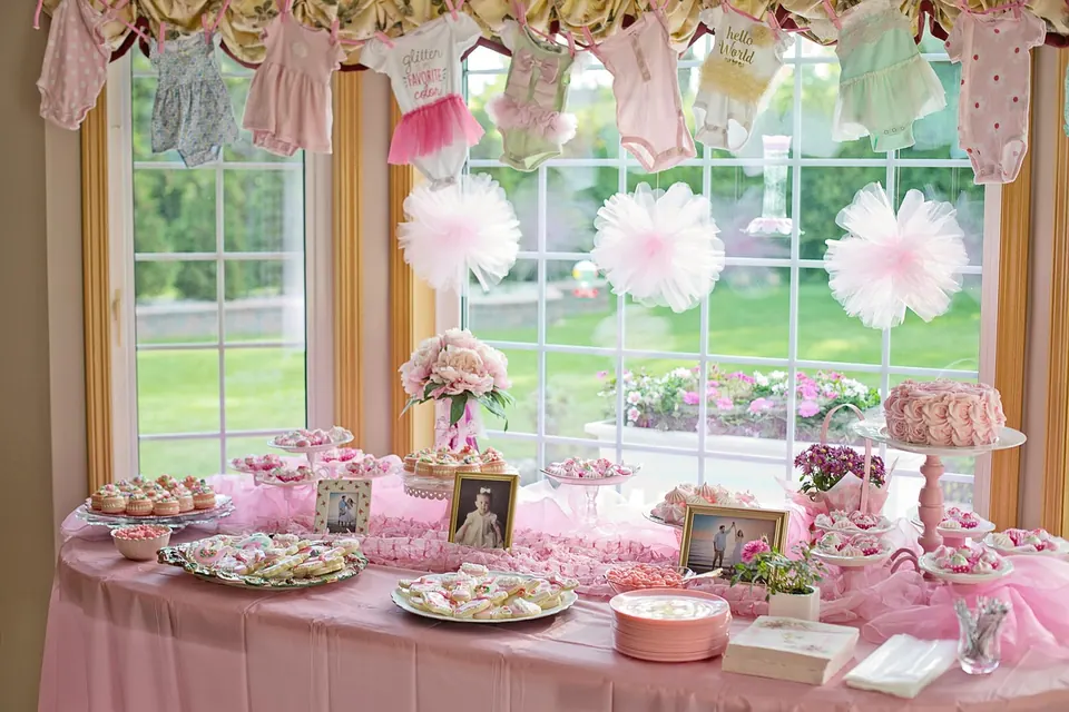 Table de shower de bébé rose avec desserts, pompons en tulle et petits vêtements suspendus à une corde devant la fenêtre